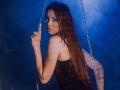 AgataRyili with long brown hair, wearing a black top, giving a middle finger, sitting on a swing with chains, blue background, looking over shoulder with a serious expression.