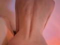 Naked back of SweetClaraa, showing slim, pale skin with a spine, small moles, and a subtle curve at the ass. Soft pink and orange lighting.