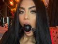 RavennaDarcy has long black hair, brown eyes, and a nose ring. She's wearing a black ball gag in her mouth with a metal ring. Her lips are slightly parted.