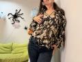 Tilliana, com uma blusa floral marrom, calças jeans escuras, a mão no peito, apoiada na parede, a cabeça inclinada para trás, cabelos castanhos longos, unhas cor-de-rosa e uma decoração de parede em formato de aranha preta..
