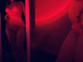 Tisia stands in red-lit room, wearing sheer pink lingerie that exposes her tits and pussy. Reflection in mirror shows her curvy ass and tattooed belly.