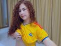 IrisDalton with curly red hair, lifting her yellow soccer shirt to expose her small, perky tits, wearing blue trim, standing in front of beige curtains.