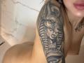 AuraXo's tattooed shoulder featuring a black-and-gray Egyptian Anubis with a fierce expression, holding a staff, and striped headpiece, with visible breasts and a partial view of her lips.