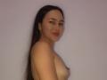 Topless MilaHans with long dark hair, smiling, showing small tits and a nipple, standing against a plain background. She's got light skin and red lipstick.