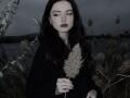 LanoraLinander with pale skin, dark lipstick, and black hair holding a fluffy cattail in a dark, moody field, wearing a black cloak. Her tits are small and covered.