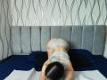 AbbyHillsI on all fours, wearing white lace lingerie, with her back to the camera, showing her ass and back tattoo. She's on a bed with dark blue sheets.