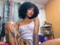 CinthiaWalker with curly black hair, wearing a checkered dress, sitting on a bed, showing tattoos on her thigh and arm, with a small nose ring and necklace.