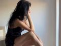 Nalini, con lunghi capelli neri, seduta, indossa un abito nero, con le gambe incrociate, le braccia appoggiate sulle ginocchia, la testa bassa, che guarda di lato, in una stanza con una luce soffusa..
