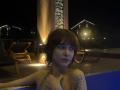 ErynAttridge with wet, shoulder-length brown hair, gazing forward, hand on chest, tattoo on left shoulder, seated in pool at night, dimly lit, wooden deck, chairs, house lights in background.