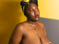 SkylerNaomi, a dark-skinned woman with red lipstick, gold hoop earrings, and a black yellow-patterned headwrap, topless, holding her tits, wearing a gold necklace, against a yellow-gray background.