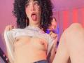 LaurenEvie69 with curly black hair, tongue out, lifting a shirt to expose small, perky tits with light brown nipples. She's sitting, legs apart, in a pink-lit room.