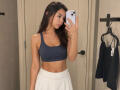 EmeraldCrivaro with long dark hair, wearing a blue sports bra, showing her small tits, and a white skirt, taking a mirror selfie with a white phone.