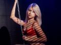 DataSley with blonde hair, red bra, holding a chain, shadow stripes on skin, looking at camera, silver necklace, slight smile, confident, sexy, attractive, soft lighting.