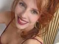 Khharha with red hair, green eyes, and pink lipstick smiling at the camera, wearing a purple top and gold hoop earrings, showing her tits and a cute face.
