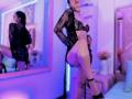 MelisaDaltonn se tient dans une pièce éclairée de lumière violette-bleue, vêtue d'une robe en dentelle noire, qui expose ses seins et ses fesses, avec une jambe levée, révélant son vagin brillant..