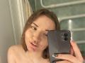 KatheyKuang with shoulder-length brown hair, holding a black phone, taking a mirror selfie, showing her nose ring and slightly open mouth, with a subtle smile.