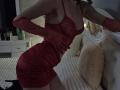 Rozalin in a red, sheer, tight dress with her tits showing, red gloves, and black high heels, leaning forward with her ass slightly raised, in a dimly lit room.