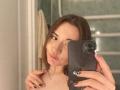 KatheyKuang with brown hair, pouty lips, holding a black phone, showing off painted nails, looking into the mirror, wearing a necklace, with a nose ring.
