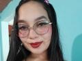 MelaniCoox has dark hair, glasses, red lipstick, and a pink hair clip. Her tits are small, and she's smiling with a slight blush on her cheeks.