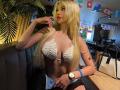ChristieGarci with long blonde hair, white lace bralette, large tits, black shorts, gold watch, finger to lips, black handbag on table, sitting in restaurant, flags above.