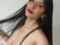 MadissonFontaine with long black hair, red lipstick, and gold earrings, topless, showing her tits, with a small mole on her right cheek, smiling at the camera.