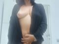 LauraSin with black shirt open, exposing her bare tits and flat stomach, wearing a silver watch, standing in a white room, her hand on her belly.