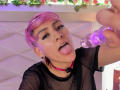 DannyRouds with pink hair, black mesh top, and pink choker, tongue out, drooling on a purple glass dildo. Earrings visible, eyelashes long, looking up at the camera.