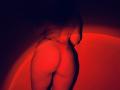 Tisia's ass, glowing red, in a sheer red thong, standing with her back to the camera, right hand on her side, shadows and light creating a sensual, provocative effect.