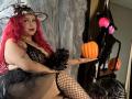 Estefy69 with red curly hair, black lace dress, fishnet stockings, holding a pumpkin, wearing a witch hat, showing a tattoo on her arm, sitting with a confident expression.