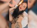 MartinaRey with long black hair, red lipstick, wearing a striped black and beige bodysuit, showing her tattooed arm and sitting on a bed with white pillows.