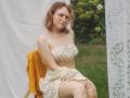 TeresaBirt with pale skin, curly red hair, glasses, small boobs, wearing a floral crop top and skirt, sitting on a chair with a yellow shirt, white lace curtain.