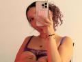 ValeryScot taking a selfie, holding her phone with one hand, showing her purple, see-through bra that barely covers her tits, with curly purple hair, and a necklace.