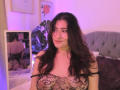 ElyRoss with long dark hair, smiling, wearing a black lace see-through top, exposing her tits, in a bedroom with a mirror reflecting her back. Soft purple lighting.