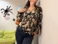 Tilliana, vestindo uma blusa floral marrom, calças jeans escuras, com as mãos no peito, encostada na parede, exibindo os seios, com uma decoração de aranha ao fundo..