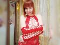 LaylaHottyX with red bob, wearing a red, fishnet, cut-out dress, arms crossed, showing her tits, and a sly smile, standing in a softly lit room.