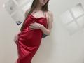 Nesie in a red satin dress, holding her hand over her pussy, showing her tits, long brown hair, white wall with square cutouts, and a blue object in the background.