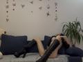 Valnes, nude, with black boots, lying on blue sofa, legs spread, arms covering tits, hand on pussy, plant in background, butterfly wall decals, dark hair, relaxed pose.