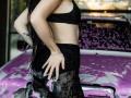 AlexLovely with long black hair, wearing a black sports bra and lace pants, standing next to a purple car covered in pink and white suds. Her hand on her ass.