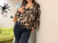 Tilliana, vestindo uma blusa marrom com estampas florais, abotoada até o peito, revelando seios pequenos, e jeans azuis, com a mão na cintura, em um quarto com decorações de Halloween, com cabelos castanhos longos..