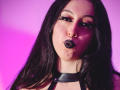 KendraSimons has dark, wavy hair, black lipstick, and a black leather choker. She's making a pouty face with pink-purple lighting highlighting her skin.
