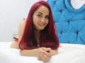 CamilaLux with red hair, smiling, laying on a bed, arms crossed, wearing a black choker, blue tufted headboard, white bed, pale skin, small tits, looking at camera.
