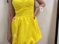 MiraMixi in a bright yellow dress with floral accents, her tits pushed up by a padded bra, hands in pockets, wearing a white flower bracelet, red nails.