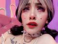 LaurenEvie69 with pink and black hair, purple eye makeup, teardrop chains, red lipstick, nose piercing, white spiked choker, ring, tattooed chest, and pigtails, showing a sexy face.