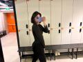 HeikeCarchi, black long-sleeve top, black pants, short dark hair, holding phone, mirror selfie, locker room, black bench, white and orange lockers, white sneakers on bench.