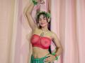 Yurimy with brown skin, green headband, red lace top revealing her tits, green skirt, arm raised, smiling, pink curtains, green wristbands, red earrings, tattoo on chest.