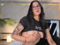 ViioletMoon, with glasses, black shirt, and tattoos, exposes her tits by cupping them with one hand, pink lips, long black hair, red bracelet, and gray pants.