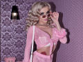 MademoiselleBella with blonde curls, wearing pink sheer lingerie, holding pink sunglasses, showing big tits, and a pink fur-sleeved top, standing in a purple, patterned room.