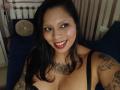 Chili, met donker haar, rode lippenstift en tatoeages op haar borst en armen, glimlacht naar de camera, draagt zwarte kant lingerie en laat haar borsten zien..