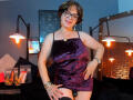 SamantaMature69 in a purple velvet dress, black stockings, glasses, smiling, hand on thigh, exposing black butt plug, in a dimly lit room with hanging lights.