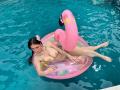 MaddyCarina with large tits, lying on a pink inflatable flamingo in a blue pool, wearing a silver chain necklace, and a flower in her black hair, legs spread.